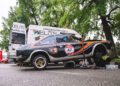 A black and blue race car with the number 47 is raised on a jack next to a white service van under tall trees, with tools and equipment on the ground during the Modena Cento Ore 25th Anniversary, held Oct. 5-10.