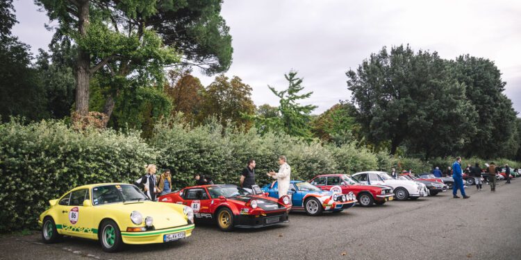 A row of classic sports cars parked along a street during the 25th Anniversary Modena Cento Ore, with people standing nearby; trees and bushes in the background under a cloudy sky from Oct. 5-10.