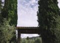 A vintage blue sports car, celebrating the 25th Anniversary of Modena Cento Ore (Oct. 5-10), is parked under a rustic stone archway between two tall cypress trees, with a cloudy sky overhead and greenery in the background.