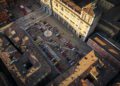 Aerial view of a city square with a fountain at the center, surrounded by parked cars and people, bordered by historic buildings in sunlight during the Modena Cento Ore 25th Anniversary, Oct. 5-10.