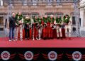 A group of people in racing suits and laurel wreaths stand on a red stage for an award ceremony at the Modena Cento Ore 25th Anniversary, held in front of a historic building from Oct. 5-10.