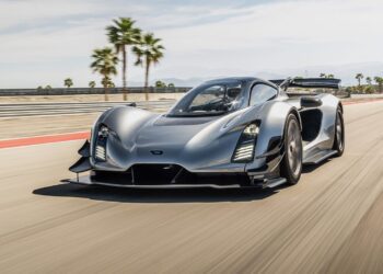 A silver American supercar drives at high speed on a racetrack with palm trees and mountains in the background.