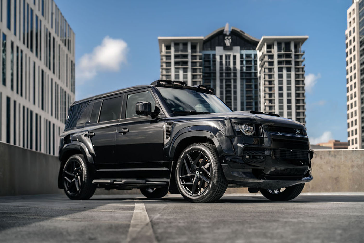 A black Land Rover Defender SUV, customized by Chelsea Truck Company, is parked in an urban setting with tall modern buildings in the background under a partly cloudy sky.