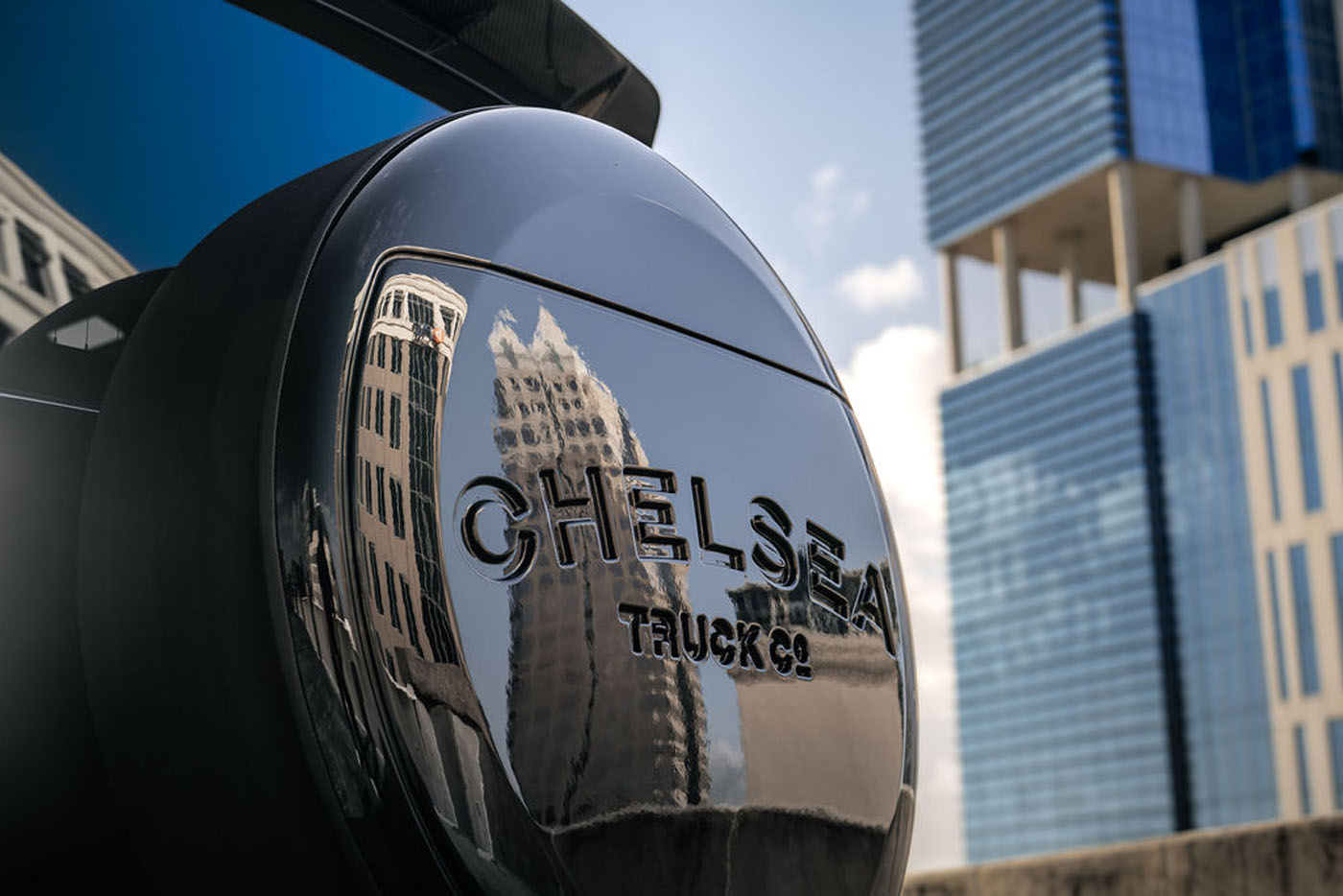 Close-up of a Chelsea Truck Company spare tire cover reflecting nearby skyscrapers, with modern glass buildings in the background—a striking touch for those who appreciate Bespoke New Defenders and urban automotive elegance.