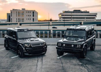 Two black SUVs—possibly Bespoke New Defenders—are parked on a rooftop parking lot at sunset, with multi-story buildings and a highway in the background.