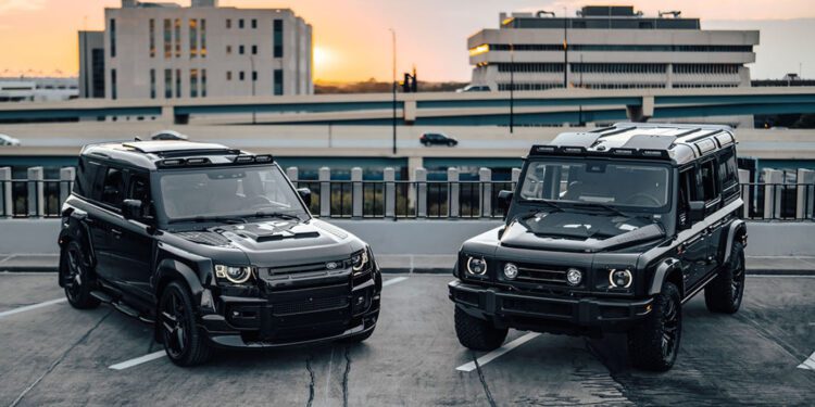 Two black SUVs—possibly Bespoke New Defenders—are parked on a rooftop parking lot at sunset, with multi-story buildings and a highway in the background.