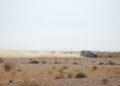 A Defender speeds across the dry, rocky Sahara Desert, kicking up a trail of dust behind it under an overcast sky—a scene worthy of the Dakar Rally.
