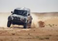 A black Defender speeds across the Sahara Desert, kicking up dust and sand under its tires as it races through the rugged terrain like a scene from the Dakar Rally.