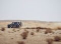 A black Defender powers swiftly across the vast Sahara Desert, kicking up a trail of dust reminiscent of the Dakar Rally.