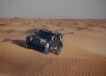 A black Dakar Rally Car ascends a sand dune in the vast Sahara Desert, set against a clear sky.