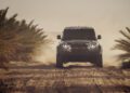 A black Defender speeds along a sandy, dirt road between palm trees, kicking up dust like it’s competing in the Dakar Rally across a Sahara Desert landscape.