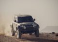 A black four-door SUV labeled "Defender" drives off-road on a dusty, rocky terrain in true Defender Tests style, raising clouds of dust as it speeds across the landscape reminiscent of the Dakar Rally.
