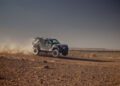 A camouflage-patterned Defender SUV drives at high speed across the rocky Sahara Desert, kicking up dust behind it under a clear sky—reminiscent of the thrilling Dakar Rally.