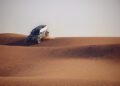A black Defender SUV drives down a sand dune, kicking up dust in the vast Sahara Desert, with rippled patterns on the sand evoking scenes from the Dakar Rally.