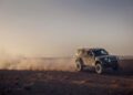 A rugged Defender SUV speeds across the barren Sahara Desert, kicking up a large cloud of dust behind it under a hazy sky.