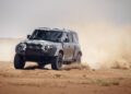 A black Land Rover Defender drives off-road on sandy terrain, kicking up dust as it powers through the Sahara Desert landscape.