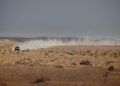A Defender drives across a dry, rocky desert landscape, kicking up a long trail of dust under the clear Sahara Desert sky.