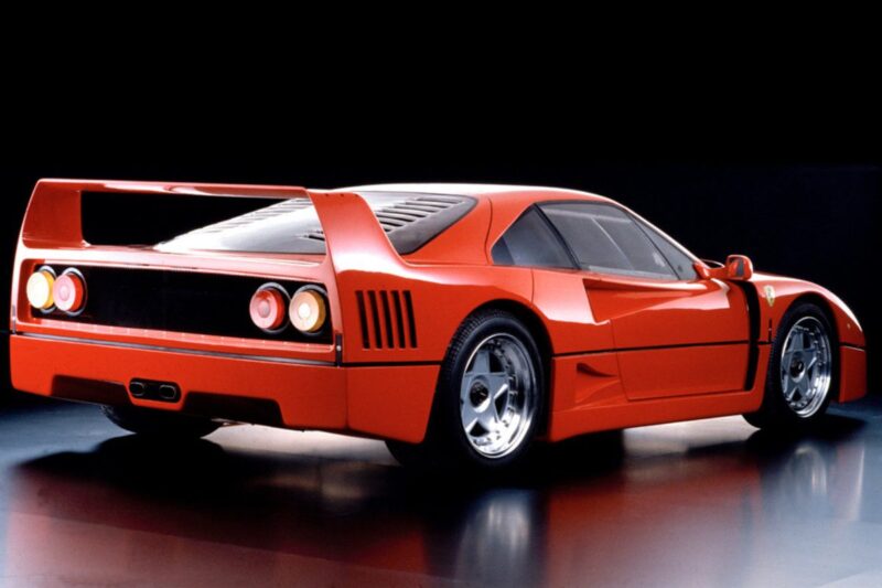 A red sports car with a large rear wing, circular tail lights, and aerodynamic design, viewed from the rear-side against a dark background—perfect for a Comparison: Ferrari F40 Vs Ferrari F50.
