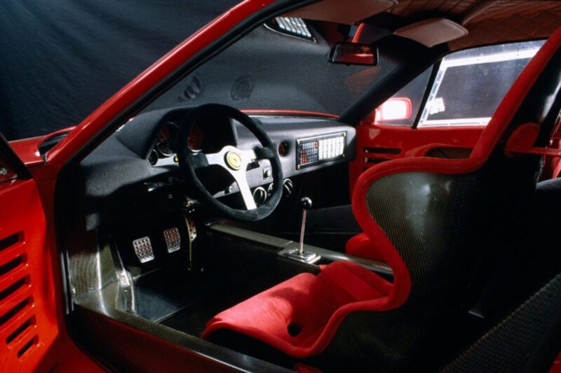 The interior of a red sports car, reminiscent of the iconic Ferrari F40 vs Ferrari F50 comparison, features a racing seat, manual gear shift, three pedals, and a steering wheel with a Ferrari emblem.