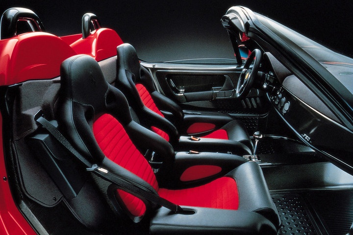 Interior of a sports car featuring black seats with red center inserts, minimalist dashboard, and visible exposed metal details—evoking the raw, purposeful design seen in icons like the Ferrari F40 Vs Ferrari F50. Viewed from the passenger side.