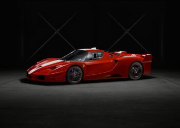 A red Ferrari FXX sports car from the exclusive XX Program is parked indoors on a dark concrete floor, illuminated against a black background with minimal lighting.