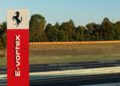 A red and white sign with the Ferrari logo and the words “E-vortex” stands beside the Ferrari test track, with trees in the background.