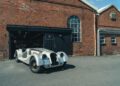 A vintage, unfinished Morgan Plus Four sports car is parked outside a brick garage building with large arched windows on a sunny day.