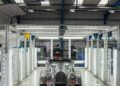A silver Morgan Plus Four sports car, celebrating its 75th Anniversary, is parked in a brightly lit, industrial workshop or factory setting.