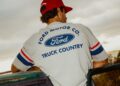 A person wearing a red cap and a gray Ford Motor Co. "Truck Country" shirt from the Limited Edition Collection leans on a green vehicle, viewed from behind.