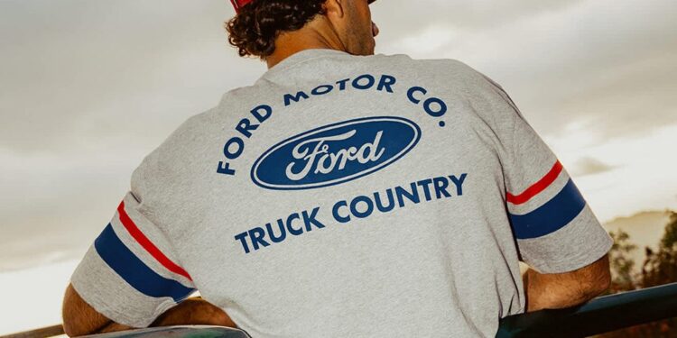 A person wearing a red cap and a gray Ford Motor Co. "Truck Country" shirt from the Limited Edition Collection leans on a green vehicle, viewed from behind.