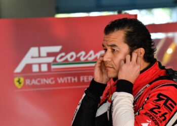 A race car driver in a red racing suit stands in front of an AF Corse Ferrari sign, covering his ears with his hands, as the duPont REGISTRY Group brings luxury and excitement to the racing world.