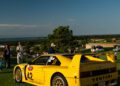 A yellow Venturi sports car with the number 42 is parked on a grassy field at The Bridge IX, capturing the spirit of automotive art; people and cars are visible in the background under a clear sky.