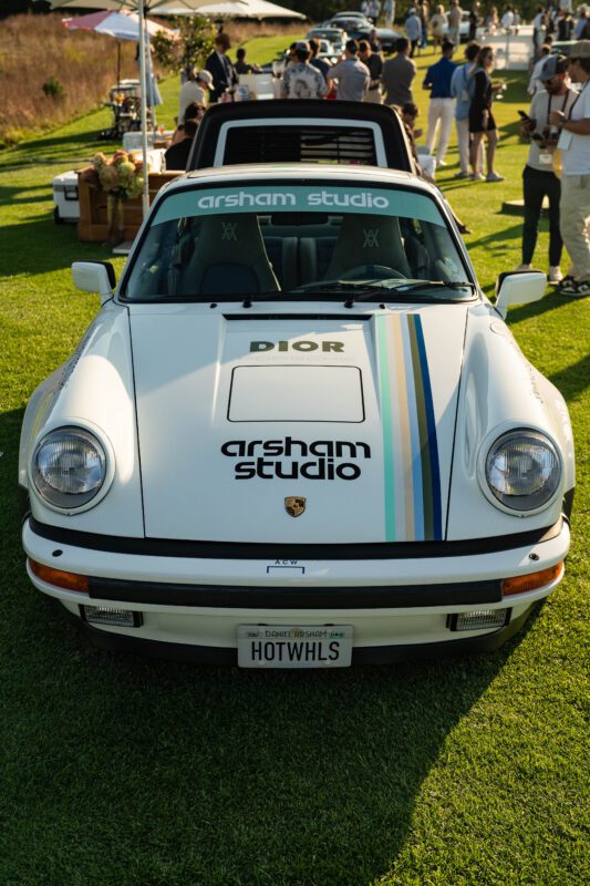 A white Porsche with "Dior" and "arsham studio" branding, colorful racing stripes, and a "HOTWHLS" license plate embodies Automotive Art at The Bridge IX, parked on grass at an exclusive outdoor event with people in the background.