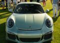 A silver Porsche sports car, a true piece of automotive art, is parked on a grassy field surrounded by people at an exclusive outdoor event.