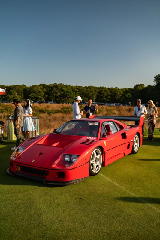 A red Ferrari F40, a true icon of automotive art, is parked on grass while several people stand and chat in the background on a sunny day.