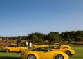 Three yellow sports cars are parked on a grassy field with their hoods open, showcasing automotive art and exclusivity, while a few people stand nearby under a clear blue sky at The Bridge IX.