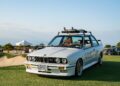 A white BMW E30 with a roof rack carrying surfboards is parked on grass under a clear blue sky, capturing the spirit of Automotive Art and exclusivity at The Bridge IX.