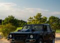 A black off-road SUV is parked on grass near sand and trees under a clear sky, capturing the essence of Exclusivity and true Automotive Art.