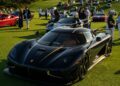 A black Koenigsegg sports car, a true piece of automotive art, is displayed on a grassy field among other luxury cars and a crowd of people at an outdoor car show.