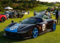 A black sports car with blue wheels and the number 23 on its hood and side is parked on a grassy field among other luxury cars and people, capturing the spirit of exclusivity at The Bridge IX—an event where automotive art takes center stage.