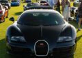 A black Bugatti sports car, a true piece of automotive art, is parked on a grassy field among other luxury cars, with people walking and standing nearby in the afternoon sunlight.
