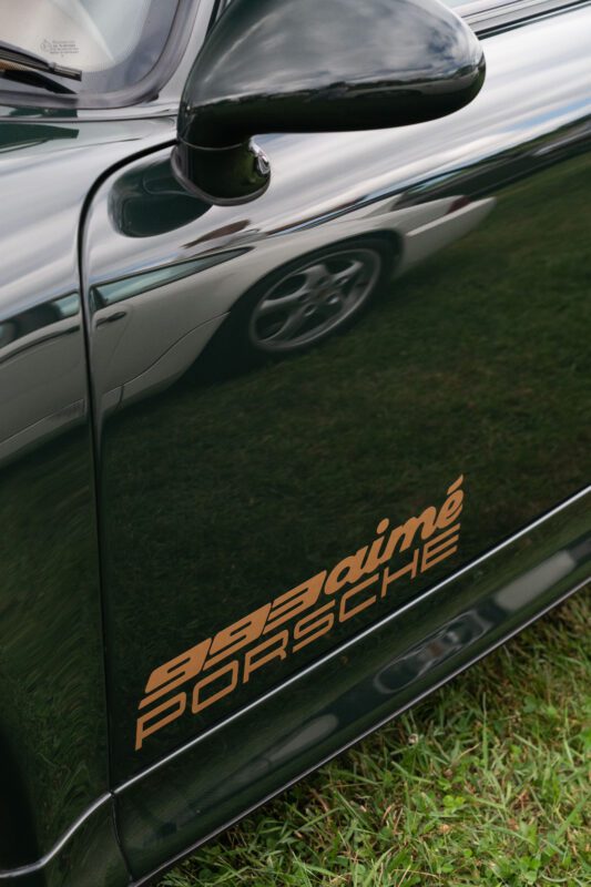 Close-up of a black Porsche car door with gold "993 aimé PORSCHE" lettering, reflecting grass and part of a nearby vehicle—an example of Automotive Art spotted at The Bridge IX, celebrating exclusivity and style.