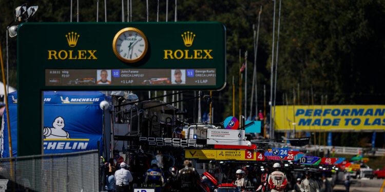 Racing teams and cars are stationed in the pit lane under a large Rolex clock and signage at Petit Le Mans, capturing the excitement of the motorsport event.