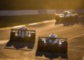 Three prototype race cars drive along a sunlit Petit Le Mans racetrack, casting long shadows in the golden light, offering a glimpse behind the scenes of this iconic motorsport event.
