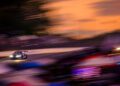 A race car with headlights on speeds along the Petit Le Mans track at sunset, with motion blur and vibrant colors in the sky and crowd, capturing the excitement of this iconic Rolex event.