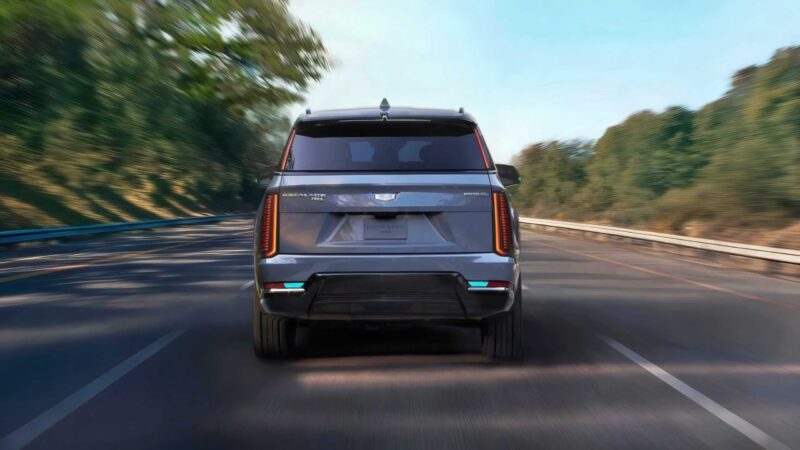 Rear view of a silver Cadillac Escalade SUV driving on a highway surrounded by trees, with motion blur indicating speed—symbolizing GM’s Next Big Leap: Conversational AI in 2026 and Eyes-Off Driving from 2028.