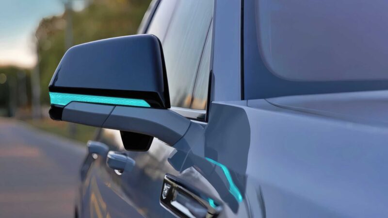 Close-up of a modern car's side mirror with an illuminated blue indicator, hinting at GM’s Next Big Leap: Conversational AI in 2026 and future eyes-off driving from 2028, parked on a road with blurred trees in the background.