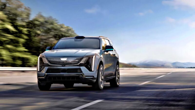 A silver Cadillac SUV, exemplifying GM’s Next Big Leap, drives on an open road with blurred trees on the side and mountains visible in the background under a blue sky.