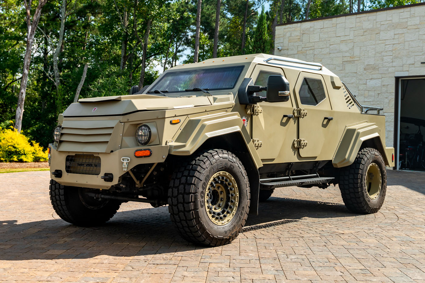 A beige street-legal armored vehicle with off-road tires is parked on a stone driveway near a modern building, surrounded by greenery.