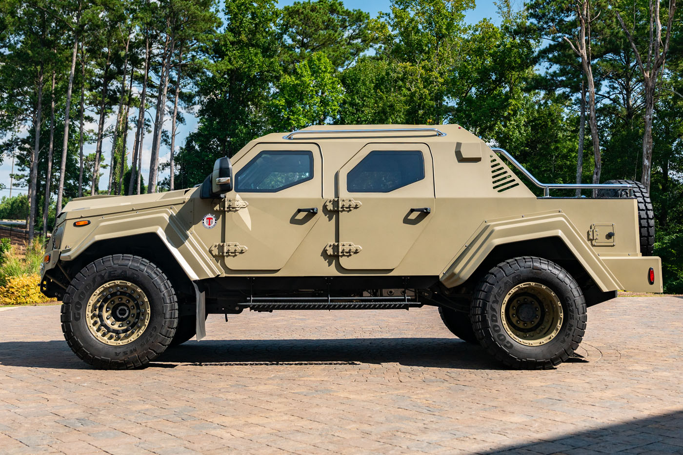 A tan-colored, armored Terradyne Gurkha with rugged tires is parked on a paved surface, surrounded by trees and greenery—this impressive street-legal vehicle is now for sale.